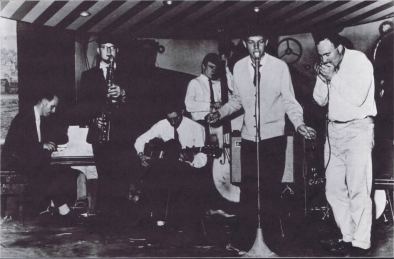 Um Mick Jagger adolescente canta no Blues Incorporated, com Jack Bruce ao fundo no baixo acústico.