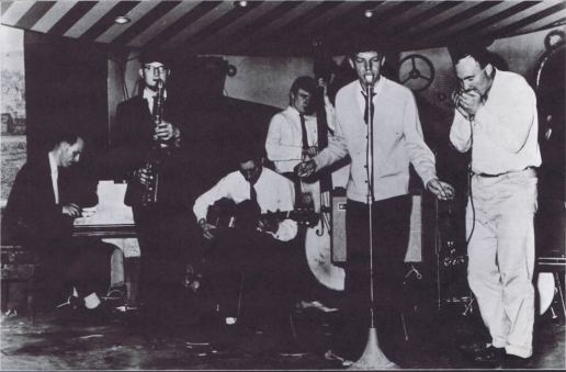 Um Mick Jagger adolescente canta no Blues Incorporated, com Jack Bruce ao fundo no baixo acústico.